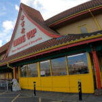 イギリスのアジア食材スーパーWing Yipの店舗外観。赤と黄色の看板が建物に掲げられ、駐車場とショッピングカートが見える。