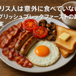 イギリスの伝統的な朝食・イングリッシュブレックファーストの写真。ベーコン、卵、ソーセージ、トースト、トマト、紅茶が木のテーブルに並び、記事タイトル『イギリス人は意外に食べていない？イングリッシュブレックファーストの起源と真実』が白文字で添えられている。